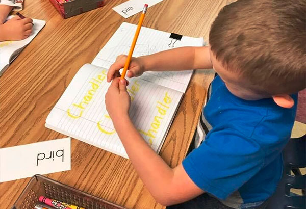 A preschool kid boy wearing blue shirt writing his name on a notebook at a Christian Preschool & Daycare Serving Loganville, GA