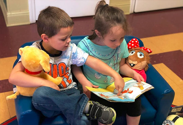 Two young boy and girl enjoying reading a storytelling book while sitting on a small foam blue sofa beside them a stuff toy at a Christian Preschool & Daycare Serving Loganville, GA