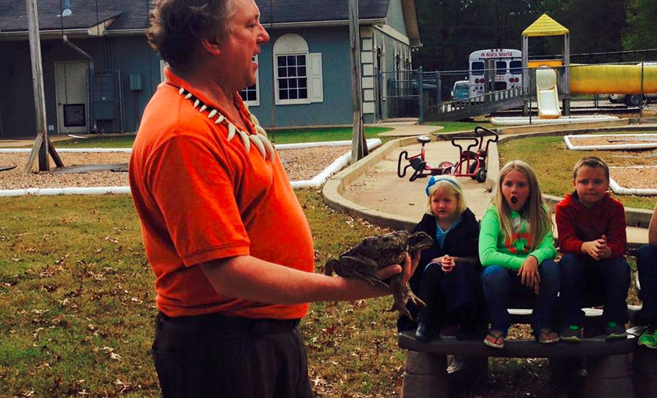 Teacher holding a big frog on his hands showing a group young kids getting amazed Group of young kids enjoying at the stadium wearing red school themed shirt at a Christian Preschool & Daycare Serving Loganville, GA