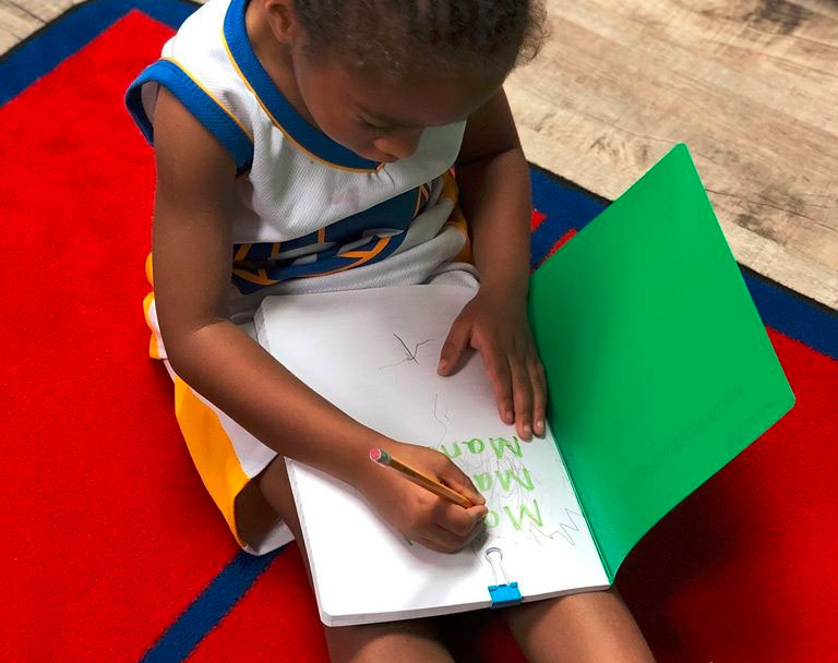 An African american preschool kid writing name on a notebook at a Christian Preschool & Daycare Serving Loganville, GA